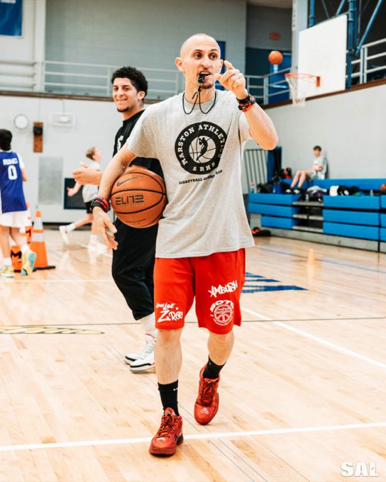 Coach Roman, whistle in his mouth and a ball under his arm, directing players at the Marston Athletics and RH2 summer camp